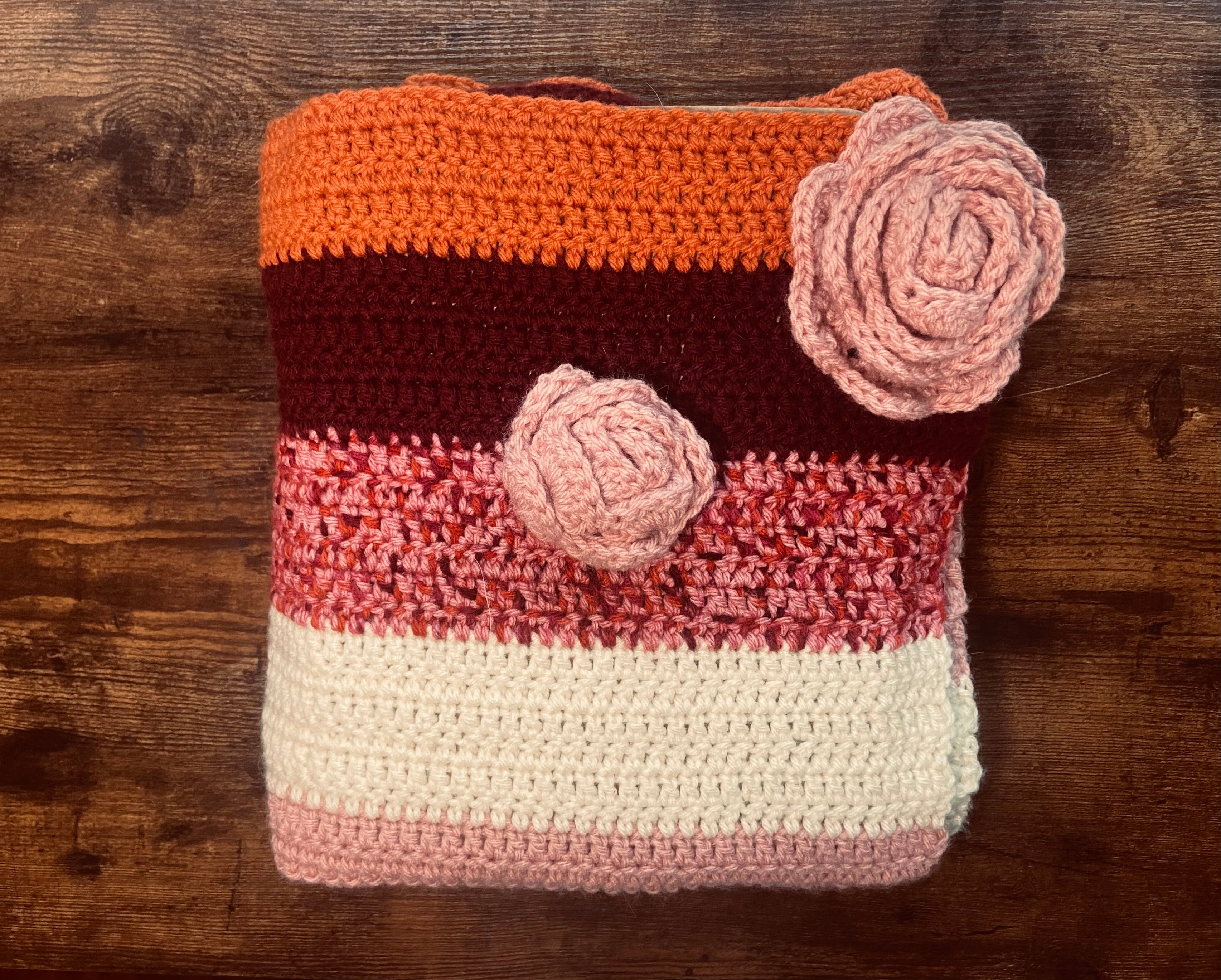 folded crochet blanket with stripes of orange, burgundy, pink, and white and light pink crochet flowers on top