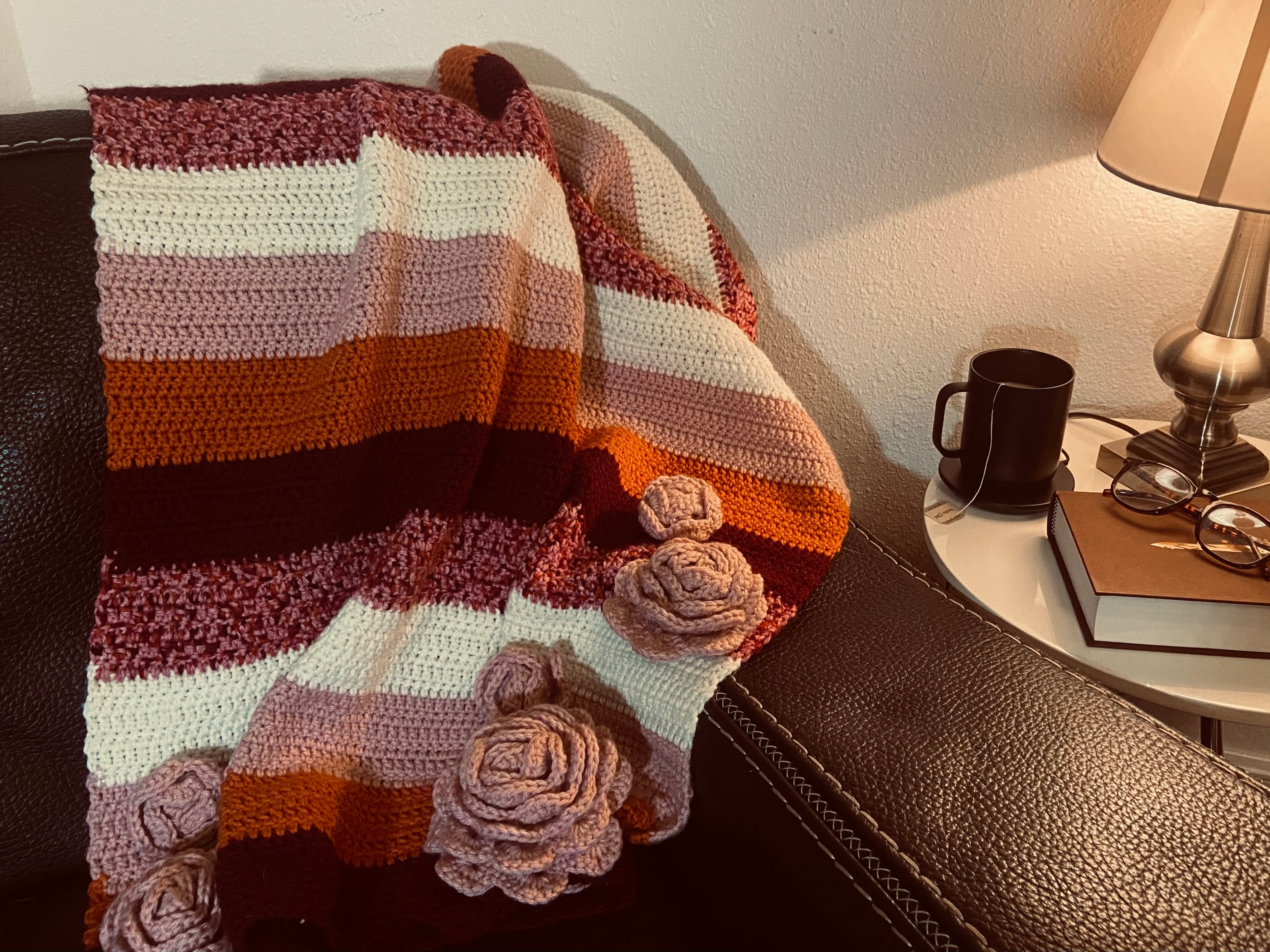 crochet blanket with stripes of orange, burgundy, pink, and white with pink crochet flowers draped over leather chair 