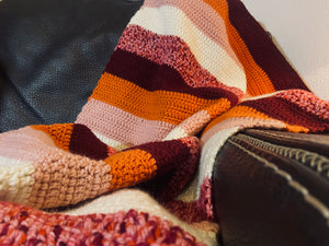crochet blanket with stripes of orange, burgundy, pink, and white draped over leather chair