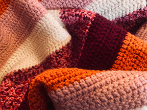 close up photo of crochet blanket with stripes of orange, burgundy, pink, and white