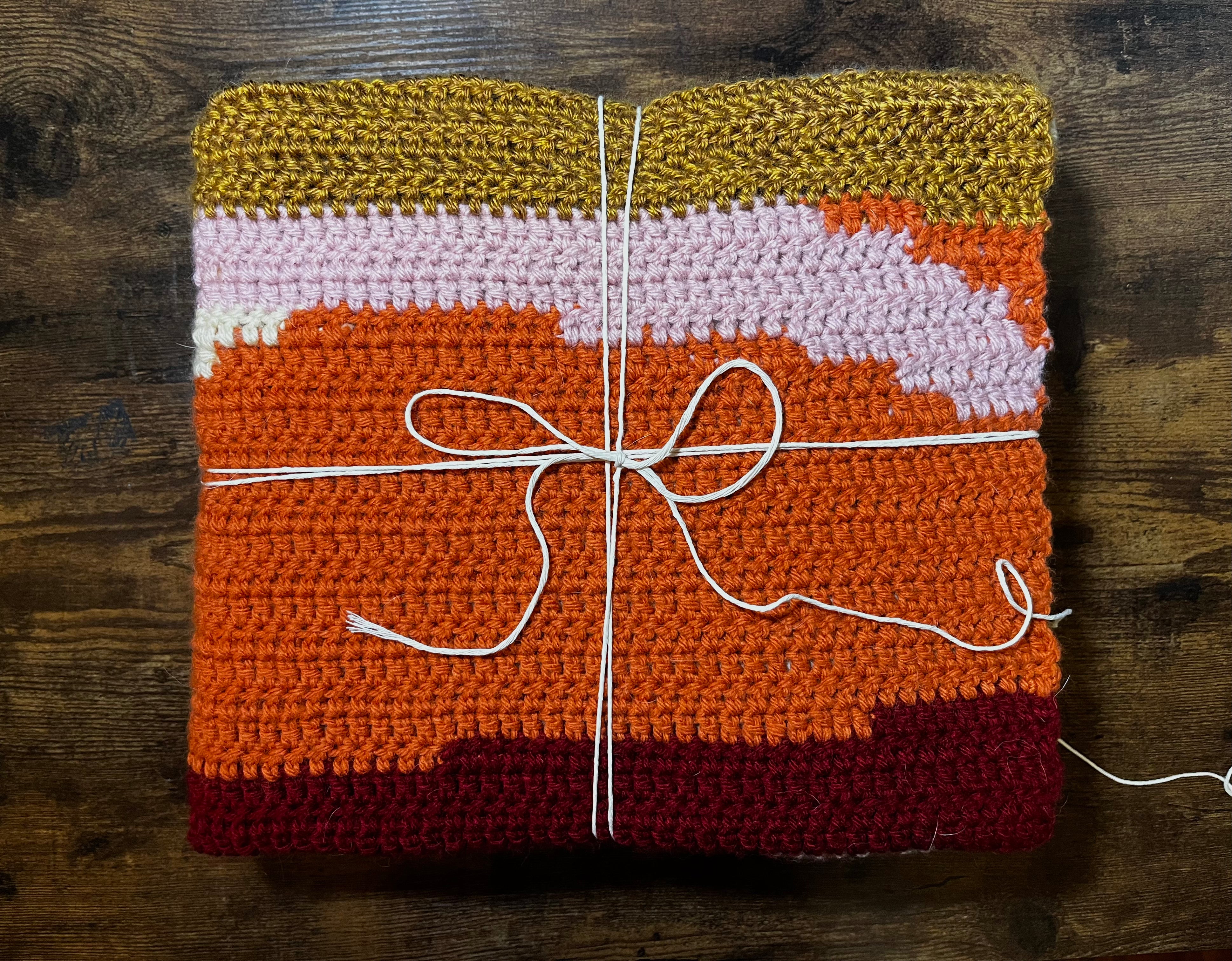 Folded crochet blanket with uneven stripes of orange, burgundy, pink, and gold.