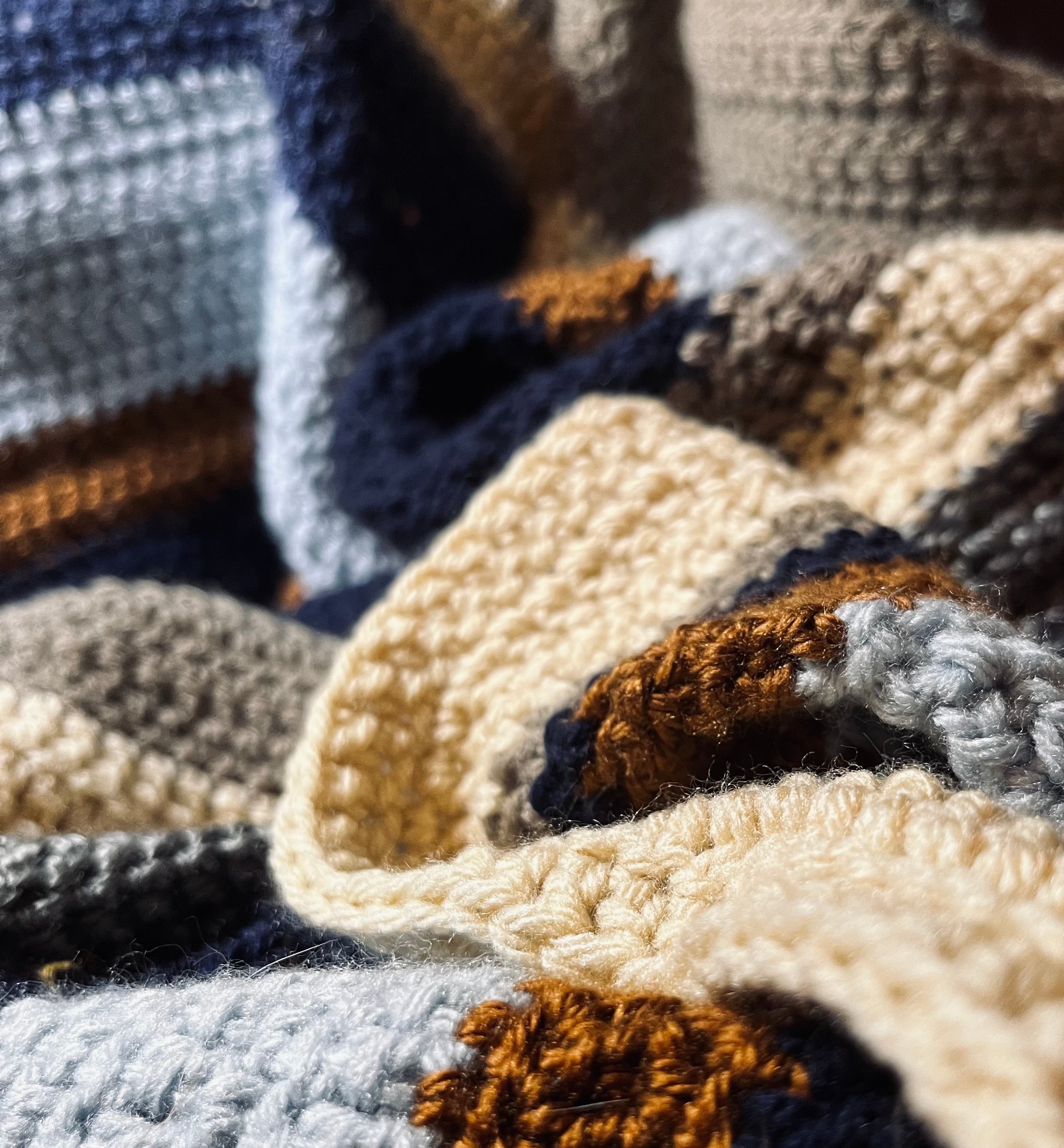 Close up photo of crochet blanket with stripes of dark blue, silver, gold, beige, tan, and light blue.