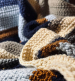 Load image into Gallery viewer, Close up photo of crochet blanket with stripes of dark blue, silver, gold, beige, tan, and light blue.
