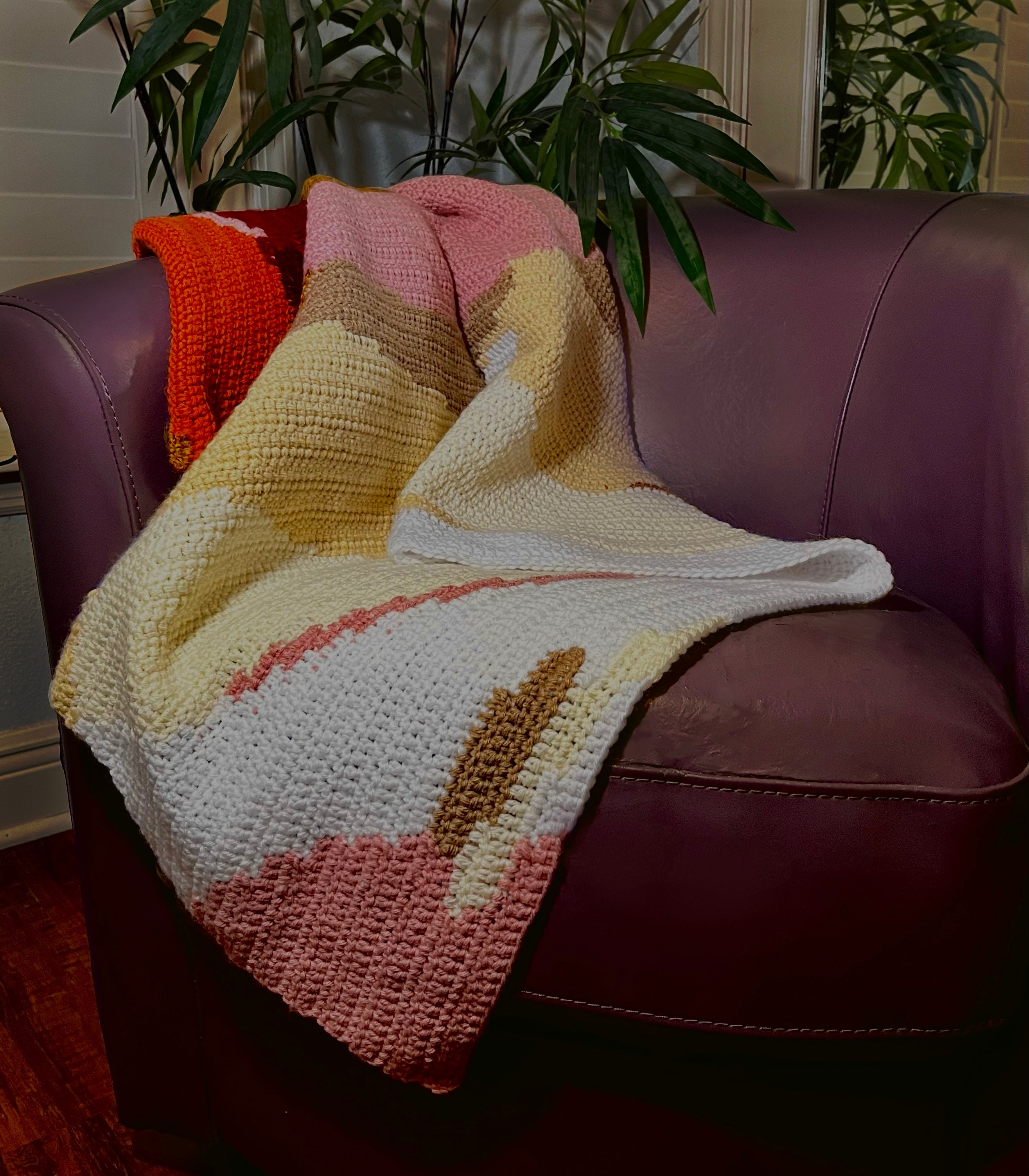 Crochet blanket with uneven stripes of orange, burgundy, pink, gold, tan, white, and off white draped over a purple leather chair.