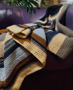 Crochet blanket with stripes of dark blue, silver, gold, beige, tan, and light blue draped over a purple leather chair.