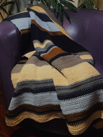 Load image into Gallery viewer, Crochet blanket with stripes of dark blue, silver, gold, beige, tan, and light blue draped over a purple leather chair.
