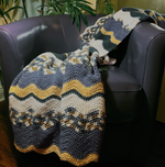 Load image into Gallery viewer, Crochet blanket with wavy stripes of light blue, white, beige, turquoise, and brown draped over a purple leather chair.
