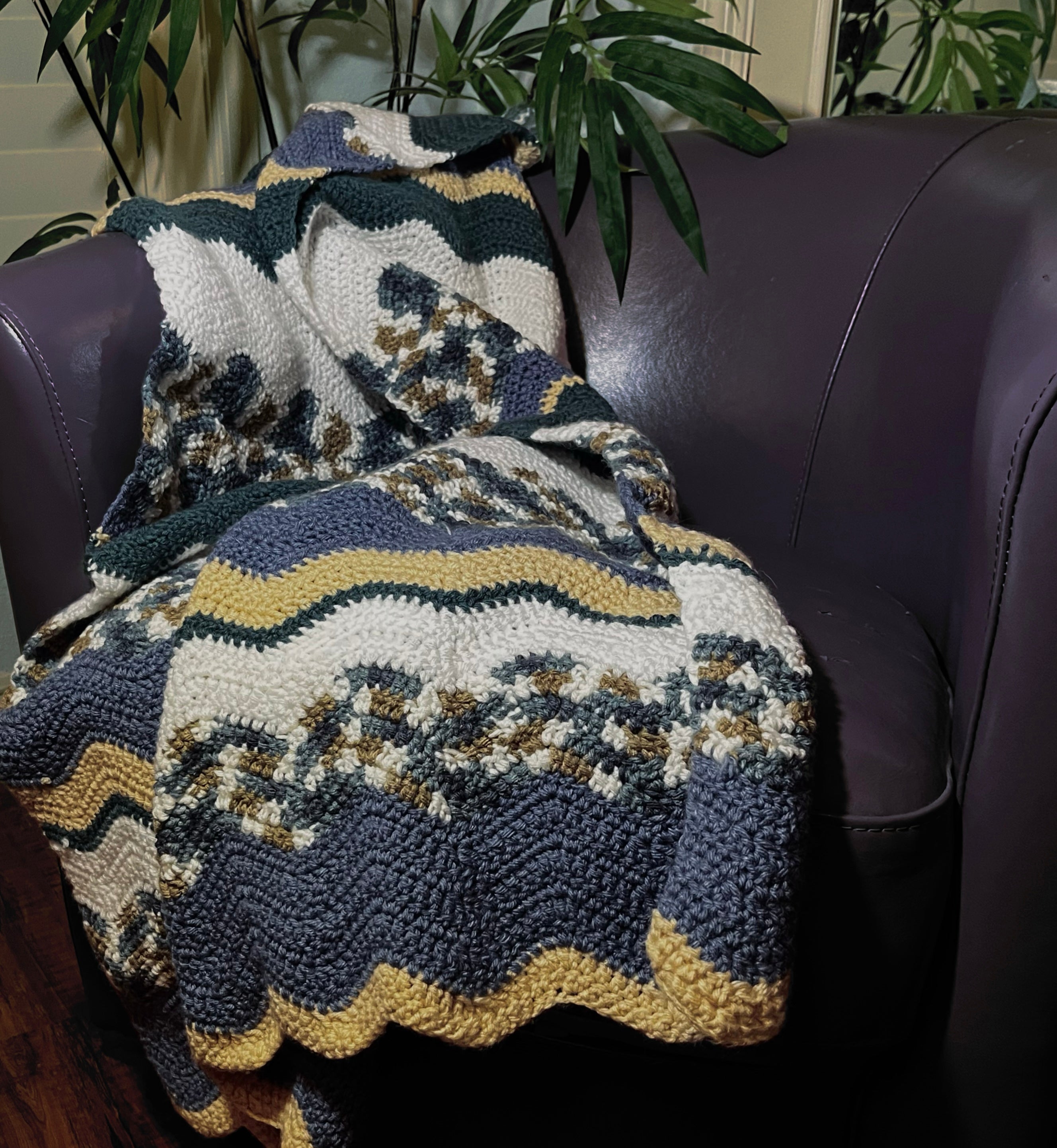 Crochet blanket with wavy stripes of light blue, white, beige, turquoise, and brown draped over a purple leather chair.