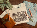 Load image into Gallery viewer, White t-shirt with black abstract wave print folded on wooden surface surrounded by blue jeans, plants, and a brown leather journal
