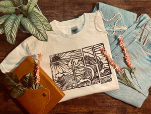 White t-shirt with black abstract wave print folded on wooden surface surrounded by blue jeans, plants, and a brown leather journal
